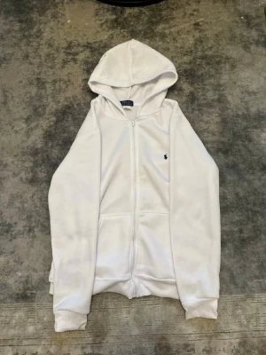 Vit zip hoodie från Polo Ralph Lauren - Snygg vit hoodie med dragkedja från Polo Ralph Lauren. Klassisk design med huva, två fickor framtill och den ikoniska lilla broderade loggan på bröstet. Tillverkad i mjukt material som känns skönt mot huden. Perfekt för en clean och avslappnad stil.