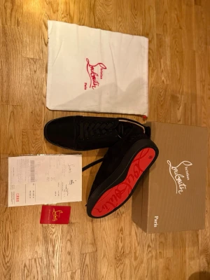 Svarta Louboutin skor - Säljer ett par svarta skor från Christian Louboutin. Skorna är en låg modell med snörning, svart ovandel och svarta skosnören. Yttersulan är röd med Christian Louboutin-logotypen. I köpet ingår original skokartong i brun/beige färg med texten ”Christian Louboutin” och ”Paris”, en vit dust bag i tyg med röd Christian Louboutin Paris-logotyp och röd dragsko, samt originalkvitto som ligger i genomskinligt plastfodral. Ett rött Christian Louboutin-kort med guldfärgad text medföljer också. Allt som s
