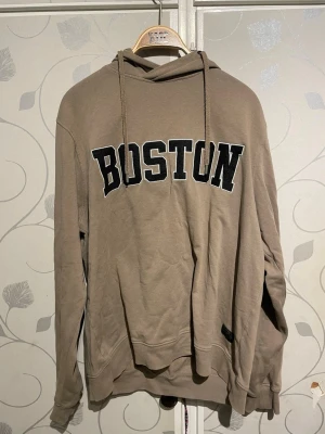 Beige hoodie med BOSTON-tryck - Snygg beige hoodie från Firefly med stort svart BOSTON-tryck på bröstet. Klassisk modell med huva och dragsnören, samt ribbade muddar vid ärmar och nederkant. Tillverkad i mjukt bomullsmaterial som är skönt att ha på sig.