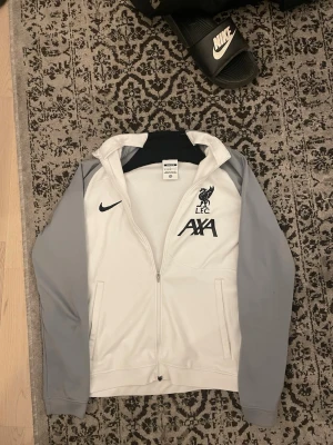Nike Liverpool zip hoodie vit/grå - Snygg zip hoodie från Nike i vitt med grå ärmar och svarta detaljer. Framsidan har Liverpool FC-logga och AXA-tryck. Två fickor framtill och huva. Perfekt för dig som gillar sportig stil och vill representera LFC. En liten del av x:et av ”axa” är lite lös