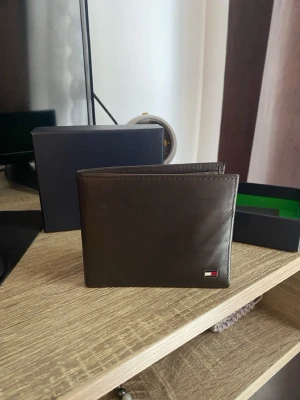 Brun läderplånbok Tommy Hilfiger - Snygg brun plånbok i äkta läder från Tommy Hilfiger. Klassisk vikbar modell med flera kortfack och diskret logga i metall på framsidan. Perfekt storlek för fickan och stilren design som passar till allt.