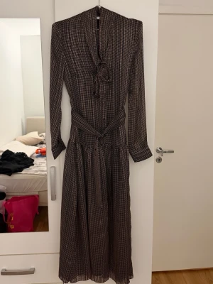 Brun långärmad klänning från ZARA - Brun maxiklänning med små rutigt mönster, långa ärmar, knytband i halsen och bälte✨Använd endast en gång - som nytt skick!