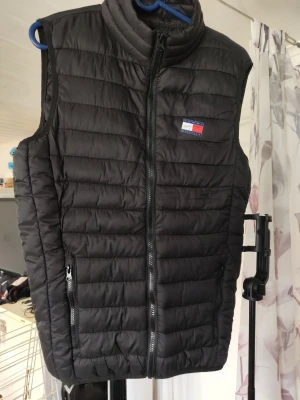 Svart väst från T Helg REA NU! - Snygg svart dunväst från T med quiltad design och hög krage. Västen har dragkedja framtill, två fickor med dragkedja och en liten logga på bröstet. Perfekt att slänga över en hoodie för en cool look. Tyvärr är de fel tryckt så loggan på bröstet är sne 