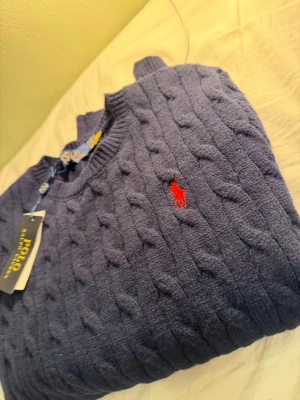 Ralph Lauren kabelstickad - Kabelstickad mörkblå tröja från Polo Ralph Lauren med klassisk röd logga broderad på bröstet. Tröjan har rund halsringning och ribbade muddar vid ärmslut och nederkant. Perfekt för dig som gillar tidlös och stilren design.