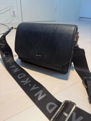 Axelväska från NA-KD - Snygg svart axelväska från NA-KD i fejkläder med gulddetaljer och bred axelrem med logga. Väskan har jättemycket utrymme och fack i den som gör den jätte praktiskt. Den har kommit till väldigt bra användning. Men det är dags att släppa taget om den då den har fått stanna hemma på senaste.🥲