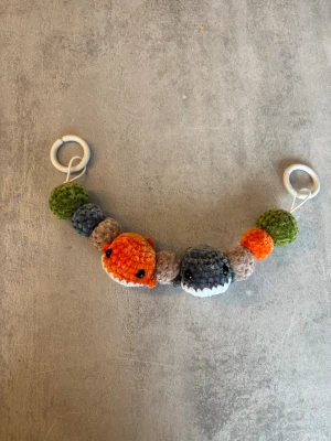 Barnvagnshänge med valar 🐳  - Charmigt barnvagnshänge i supermjukt sammetsgarn (Bohéme velvet). Hänget består av söta små valar och bollar i harmoniska naturfärger – orange, grå, beige och grön – som ger ett lugnt men lekfullt intryck. Perfekt för små nyfikna ögon 💫  ⸻  ✨ Detaljer - Ca 47 cm långt (från ring till ring) och med rundresår som gör den elastisk - Virkat i Bohéme velvet – extra mjukt och gosigt - Fästs enkelt med ringar i båda ändar (passar de flesta barnvagnar, babyskydd m.m.) - Lätt, mjukt och dekorativt 