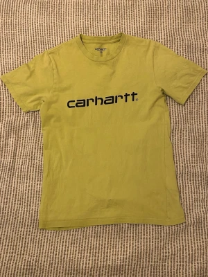 Limegrön Carhartt t-shirt med logga - Limegrön t-shirt från Carhartt med svart logotyp tryckt över bröstet. Den är lite mer åt det grönare hållet än det ser ut på bilderna. Den passar mig som är 174 ganska bra. Men både mindre och lite större personer kan ha den. Det är ett pyttelitet hål på framsidan men som inte syns från avstånd.