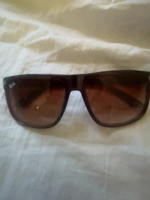 Bruna solglasögon från Ray-Ban - Snygga solglasögon från Ray-Ban med bred, brun plastbåge och tonade bruna glas. Klassisk och stilren design med Ray-Ban-logga i vitt på linsen. Perfekta för soliga dagar och ger en cool vibe till din outfit.