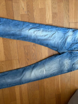 Blå slitna jeans från Nudie Jeans - Säljer ett par blå jeans från Nudie Jeans med slitningar och distressed detaljer på benen. Jeansen har klassisk femficksmodell, raka ben och en snygg tvättad look. Materialet är denim i bomull och bakfickan har Nudie Jeans signaturdetalj.