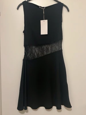 Svart klänning med spets från Zara - Snygg svart klänning från Zara med transparent spetsdetalj i midjan. Klänningen är ärmlös, har en lätt utställd kjol och är gjord i ett mjukt tyg. Spetsen ger en cool kontrast och gör plagget extra trendigt.