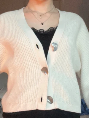 Vit ribbstickad kofta med stora knappar - Säljer en krämvit ribbstickad kofta med oversized passform och unika, stora silverfärgade knappar. Koftan har v-ringning och långa ärmar, perfekt att slänga över ett linne eller t-shirt. Materialet känns mjukt och stretchigt.