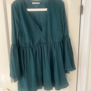 Grön omlottklänning med v-ringning - Snygg grön kortklänning med v-ringad omlottfront och långa, vida ärmar. Klänningen är aldrig använd pga för liten. Knappen (se sista bilden) har tappats.
