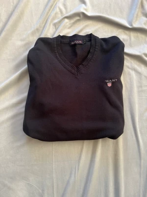 Svart stickad tröja från GANT - Klassisk svart stickad tröja från GANT med V-ringning och ribbade muddar. Tröjan har en broderad GANT-logga med sköld på bröstet. Perfekt basplagg i garderoben och passar till det mesta. Materialet känns mjukt och följsamt. Obs det står storlek S men den passar 158 unisex me ganska sliten me säg till om du vill ha flera bilder 