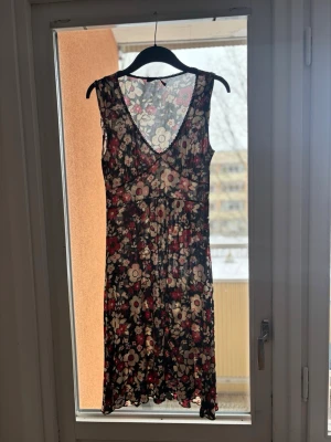 Blommig sommarklänning med v-ringning - Säljer en blommig kortklänning med v-ringning och ärmlös design från Sandro Feerone. Klänningen har ett svart tyg med vita och röda blommor, vilket ger en retrovibe. Perfekt för varma dagar och enkel att styla med sneakers eller sandaler.