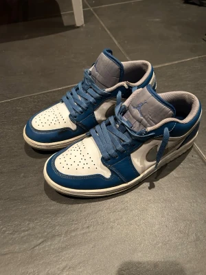 Air Jordan 1 Low blå och vita sneakers - Säljer ett par Air Jordan 1 Low sneakers i blått och vitt läder med grå detaljer. Skorna har perforerad tåbox, blå snörning och den klassiska loggan på plösen. Snygg och bekväm passform.