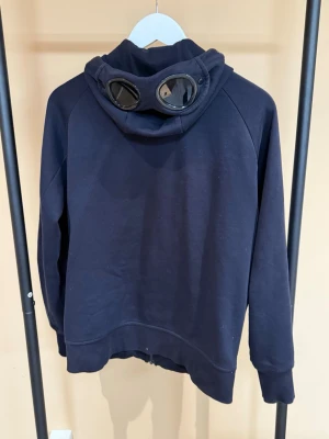 Mörkblå hoodie med glasögon i huvan - Säljer en mörkblå hoodie från C.P. Company med dragkedja framtill och två fickor med dragkedja. Huvan har de ikoniska inbyggda glasögonen som ger en unik streetwear-look. Tillverkad i mjukt bomullsmaterial och har långa ärmar med muddar.