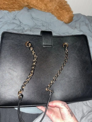 Svart handväska med kedjerem - Snygg svart handväska från Anna Field i konstläder med guldfärgade kedjeremmar och ett stilrent spänne framtill. Väskan har en rektangulär form och klassisk design som gör den enkel att matcha med olika outfits. Perfekt för dig som gillar en elegant och tidlös look.