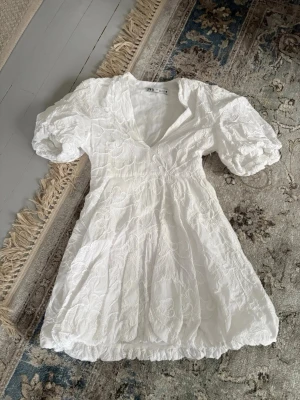 Vit studentklänning från Zara - Superfin vit kort klänning från Zara med puffärmar och v-ringning. Klänningen har broderade blommönster och en lätt volang nertill. Perfekt för studenten. Finns även dragkedja på sidan (se bild). Ingen prislapp kvar men är aldrig använd, köpt i somras.