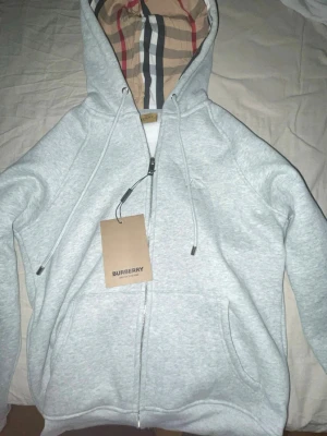 Grå zip hoodie från Burberry - Helt oanvänd och ny Snygg grå zip hoodie från Burberry med klassiskt rutigt foder i huvan och dragsko. Hoodien har fickor framtill och metalländar på snörena. Perfekt för dig som vill ha en stilren och exklusiv look med ikoniska Burberry-detaljer. Obs strl L men känns som M/S