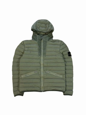 Grön pufferjacka från Stone Island - Snygg grön pufferjacka från Stone Island med huva och två snedställda fickor med dragkedja. Jackan har den klassiska Stone Island-loggan på vänster ärm och quiltad design. Perfekt för kalla dagar och riktigt trendig stil. Retail runt 9k. Passar S mer än M. Funkar nog vinter och tidig vår. 