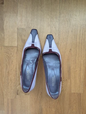 Ljuslila pumps från Sisters - Unika pumps från Sisters i ljuslila med mörklila och vinröda detaljer. Skorna har fyrkantig tå och låg klack, samt kontrastfärgade kanter. Tillverkade i konstläder med en cool retrovibe. Perfekta för dig som vill sticka ut med din stil.