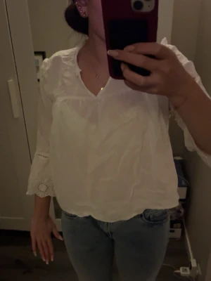 Vit blus med spetsdetaljer - Söt vit blus med v-ringning och volang vid axlarna. Ärmarna är långa och avslutas med breda spetsdetaljer och hålmönster. Blusen är gjord i ett tunt, lätt material som ger en luftig känsla. Det är en liten fläck som jag visar på en av bilderna där av priset men inget som sticker ut så.