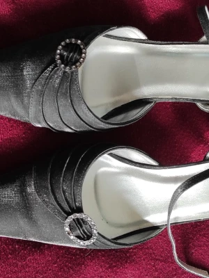 Svarta pumps med strassdetalj - Eleganta svarta pumps med spetsig tå och dekorativ strassprydd ring framtill. Skorna har en slät satinliknande yta och tunna remmar. Perfekta för dig som vill ha en klassisk och stilren look med lite extra bling.