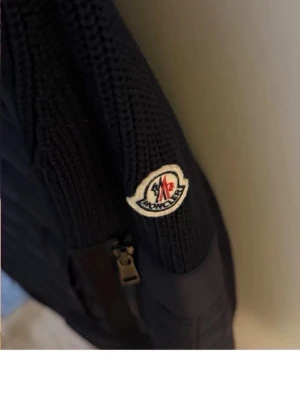Svart cardiganjacka från Moncler - Snygg svart cardiganjacka från Moncler med stickade ärmar och vadderad framsida. Jackan har dragkedja och en ikonisk Moncler-logga på ärmen. Perfekt för dig som vill ha en stilren och exklusiv look.