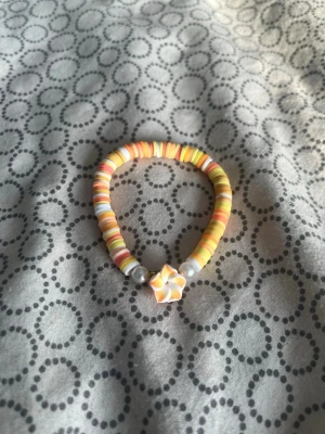 Färgglatt pärlarmband med blomma - Ett handgjort armband med platta pärlor i gula, orangea och vita toner. Armbandet har två vita pärlor och en detalj i form av en blomma i orange och vitt. Perfekt för dig som gillar färgglada accessoarer och unika detaljer.