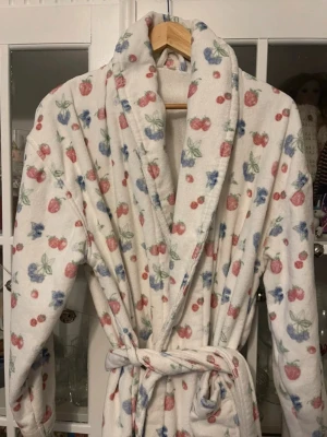 Vit morgonrock med bär från Djerf Avenue - Mysig vit morgonrock från Djerf Avenue med bälte i midjan och färgglatt bärmönster i rosa, blått och grönt. Tillverkad i mjuk frotté som känns skön mot huden. Perfekt för lata morgnar eller kvällar hemma.