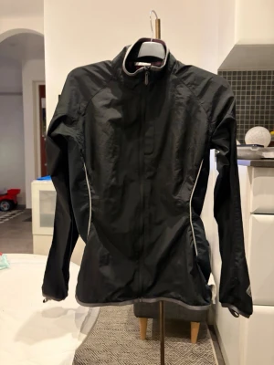 Svart vindjacka med reflexdetaljer - Snygg svart vindjacka med hög krage och dragkedja framtill. Jackan har vita reflexdetaljer längs sidorna och på ryggen, samt en praktisk ficka med dragkedja bak. Tillverkad i lätt och vindtätt material, perfekt för blåsiga dagar.