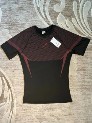 Svart träningströja från Gymshark - Snygg svart träningströja från Gymshark med röda detaljer och logga på bröstet. Tillverkad i ett stretchigt och snabbtorkande material som passar perfekt för intensiva träningspass. Kortärmad och figurnära passform för maximal rörelsefrihet.