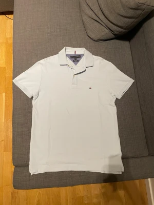 Ljusblå pikétröja från Tommy Hilfiger - Klassisk ljusblå pikétröja från Tommy Hilfiger med liten logga på bröstet. Tröjan har krage, korta ärmar och knäppning med två knappar. Perfekt till en avslappnad men ändå stilren look. Pris går att diskuteras