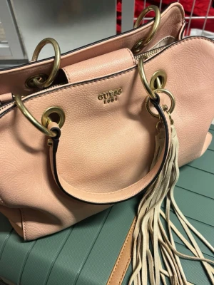Ljusrosa handväska från Guess - Snygg ljusrosa handväska från Guess med guldfärgade metalldetaljer och stora ringar. Väskan har dubbla handtag och en dekorativ tofs i mockaimitation. Tillverkad i slätt konstläder med Guess-logga i guld framtill. Perfekt för dig som gillar trendiga accessoarer.