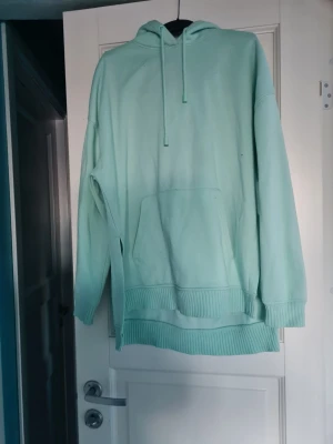 Mintgrön oversized hoodie - Snygg mintgrön hoodie med huva och dragsko. Hoodien har en stor magficka och ribbade muddar vid ärmslut och nederkant. Perfekt för en avslappnad och chill stil. Materialet känns mjukt och bekvämt, troligen bomullsblandning. Har en liten fläck på framsidan av hoodien.