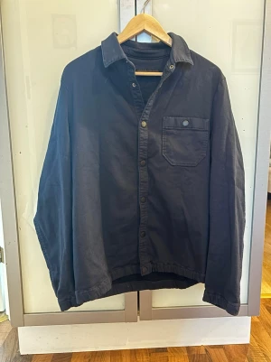 Blå overshirt från Drykorn - Stilren blå overshirt från Drykorn med klassisk krage, knappstängning framtill och en bröstficka med knapp. Tillverkad i ett mjukt bomullstyg som ger en avslappnad look. Perfekt att bära över en t-shirt för en enkel men snygg outfit. Eller bära över en schysst tröja. Perfekt nu inför våren! 