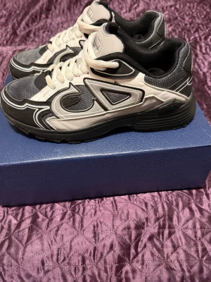  Dior b30 - Snygga Dior sneakers i svart och vitt med coola detaljer och meshpaneler. Skorna har en chunky sula och vita skosnören. Märket syns tydligt på plösen och designen är sportig med futuristiska linjer. Perfekta för dig som gillar streetwear och exklusiva märken.
