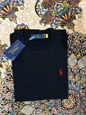 Svart t-shirt från Polo Ralph Lauren - Klassisk svart t-shirt från Polo Ralph Lauren med rund hals och det ikoniska röda broderade logotypen på bröstet. Tillverkad i mjuk bomull för en skön känsla och perfekt passform. Enkel och stilren design som funkar till allt.