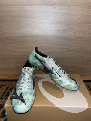 Mizuno alpha  fotbollsskor storlek 42,5 - Snygga fotbollsskor från Mizuno i vitt med skimrande detaljer och svarta loggor på sidorna. Skorna har snörning och sulor med neonfärgade dobbar för bra grepp på planen. Lätta och smidiga, perfekta för snabba rörelser och kontroll.