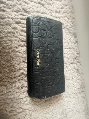Svart plånbok från Calvin Klein - Snygg svart plånbok från Calvin Klein i konstläder med präglad logotyp över hela ytan och guldfärgad metallogo framtill. Plånboken har en avlång form och stilren design som passar perfekt i väskan. Perfekt accessoar för dig som gillar klassisk och trendig stil. Köpt också i USA för $79,50 vilket är runt 800-900 kr säljer för jag inte använder men ÄLSKAR den sååå mycket! Har skråmor därför jag säljer billigt 