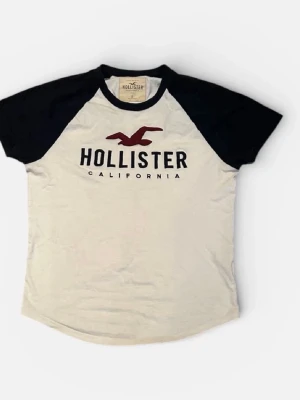 Vit och svart Hollister t-shirt - Snygg t-shirt från Hollister med svart raglanärm och vit kropp. Stor Hollister-logga och röd fågel på bröstet. Klassisk rund halsringning och mjukt bomullsmaterial gör den perfekt till vardagslooken.