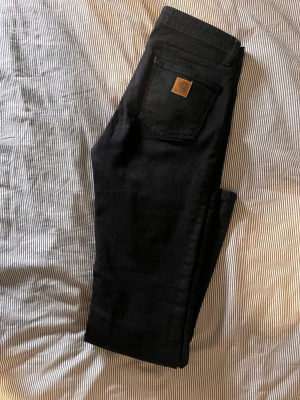Svarta jeans från Carhartt - Säljer ett par svarta jeans från Carhartt med klassisk femficksdesign och brunt Carhartt-märke på bakfickan. Jeansen har rak passform och är tillverkade i slitstarkt bomullstyg. Perfekta för dig som gillar streetwear och enkel stil.