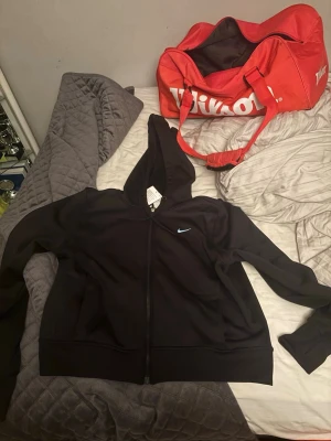 Svart Nike hoodie med dragkedja - Snygg svart hoodie från Nike med dragkedja framtill och liten vit Nike-logga på bröstet. Tillverkad i mjukt och funktionellt Therma-FIT-material som håller värmen. Huva med snörning och två fickor på sidorna. Perfekt för chill eller träning. Prislapp finns kvar!
