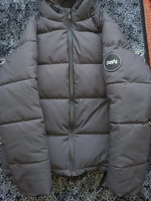 Svart pufferjacka från Supply & Demand - Snygg svart pufferjacka från Supply & demand med huva och dragkedja framtill. Jackan har ett stort runt märke på ärmen med texten 'Supply Exploration Programme Performance Pursuit'. Perfekt för kalla vinterdagar och riktigt trendig look.