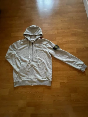 Grå zip hoodie från Stone Island - Säljer en grå zip hoodie från Stone Island med klassisk huva och dragsko. Hoodien har ribbade muddar, två fickor fram och den ikoniska Stone Island-patchen på ärmen. Tillverkad i mjuk bomullsblandning som är skön att ha på sig. Kvitto och bevis finns, kom dm