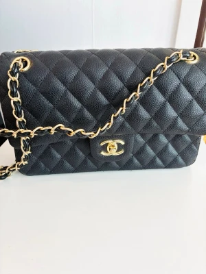 Snygg svart quiltad axelväska - Säljer en klassisk svart quiltad axelväska med guldfärgade detaljer och kedjerem. Väskan har det ikoniska CC-spännet framtill och är tillverkad i läder med struktur. Vet ej om den är äkta
