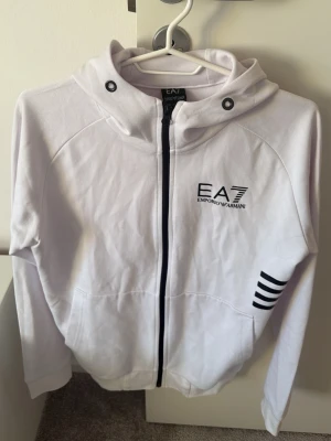 Vit EA7 hoodie med dragkedja - Snygg vit hoodie från EA7 Emporio Armani med svart dragkedja, huva och svarta detaljer. Logga på bröstet och tre svarta ränder på sidan. Tillverkad i mjukt material, perfekt för en avslappnad och trendig look.