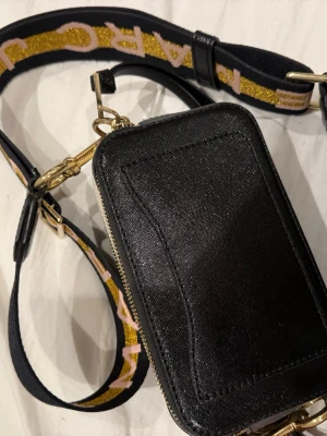 Svart axelväska Marc Jacobs - Säljer en svart axelväska från Marc Jacobs i läder med guldfärgade metalldetaljer. Väskan har en bred axelrem med glitterdetaljer och logga i gult och rosa. Praktisk dragkedja och stilren, boxig form. Perfekt för dig som vill ha en trendig och snygg accessoar.