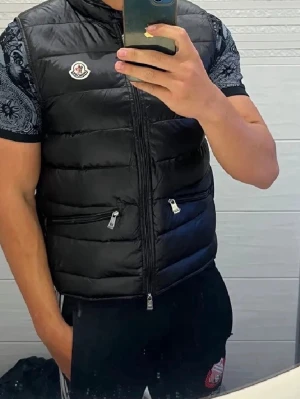 Svart dunväst från Moncler  - Snygg svart dunväst från Moncler med quiltad design och två dragkedjeförsedda fickor framtill. Västen har hög krage och Moncler-logga på bröstet. Perfekt att slänga över en t-shirt för en trendig look.
