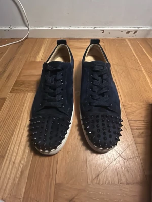 Marinblå sneakers med nitar - Snygga marinblå sneakers från Christian Louboutin med svarta nitar över tån. Skorna har en låg silhuett, svarta skosnören och vit sula. Ovandelen är i mocka och insidan är ljusbeige. Perfekta för dig som vill sticka ut. Kommer med box